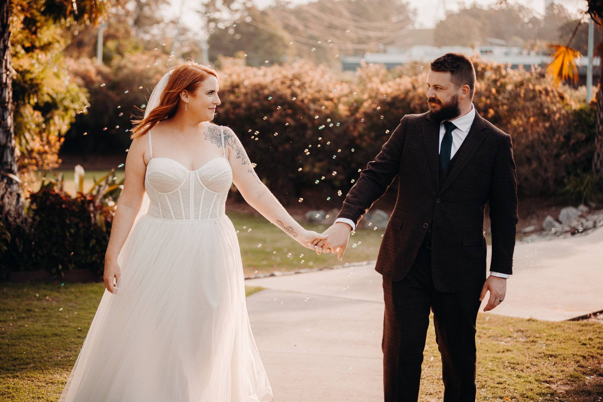 The bride Lilas in a white wedding dress and the groom Kaine in a dark suit hold hands and walk outdoors at Eatons Hill Hotel during their couple portraits session.