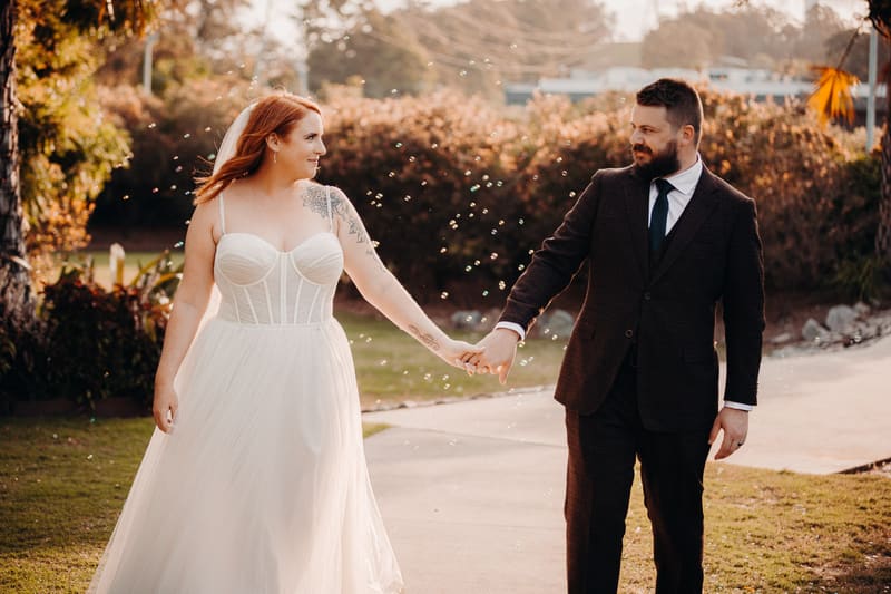 The bride Lilas in a white wedding dress and the groom Kaine in a dark suit hold hands and walk outdoors at Eatons Hill Hotel during their couple portraits session.
