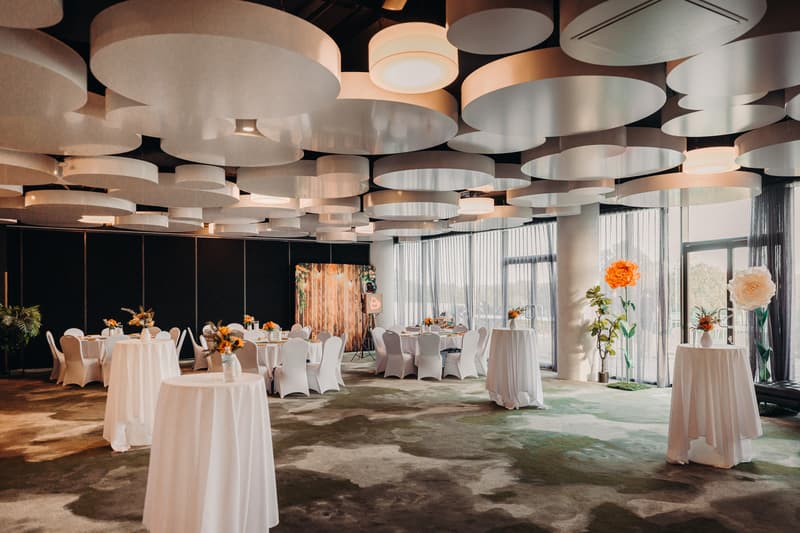 Reception room at Eatons Hill Hotel — Hills Room set with round tables and cocktail tables covered in white linens, decorated with floral centerpieces and large artificial flowers near the windows.