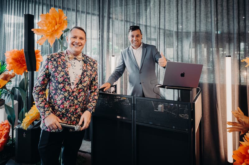 Two men stand near a DJ booth with a laptop at Eatons Hill Hotel — Hills Room, surrounded by large orange floral decorations and sheer curtains.