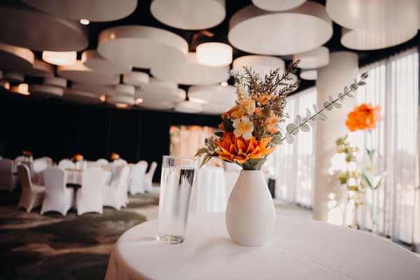 A floral centerpiece in a white vase and a glass of water on a round table at the Eatons Hill Hotel — Hills Room, with tables and chairs covered in white cloth in the background.