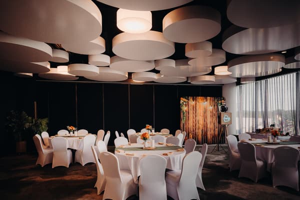 Empty reception room at Eatons Hill Hotel — Hills Room with round tables covered in white tablecloths and white chairs, each table decorated with floral centerpieces. The room features large circular ceiling lights and a photo booth setup near a window with sheer curtains.