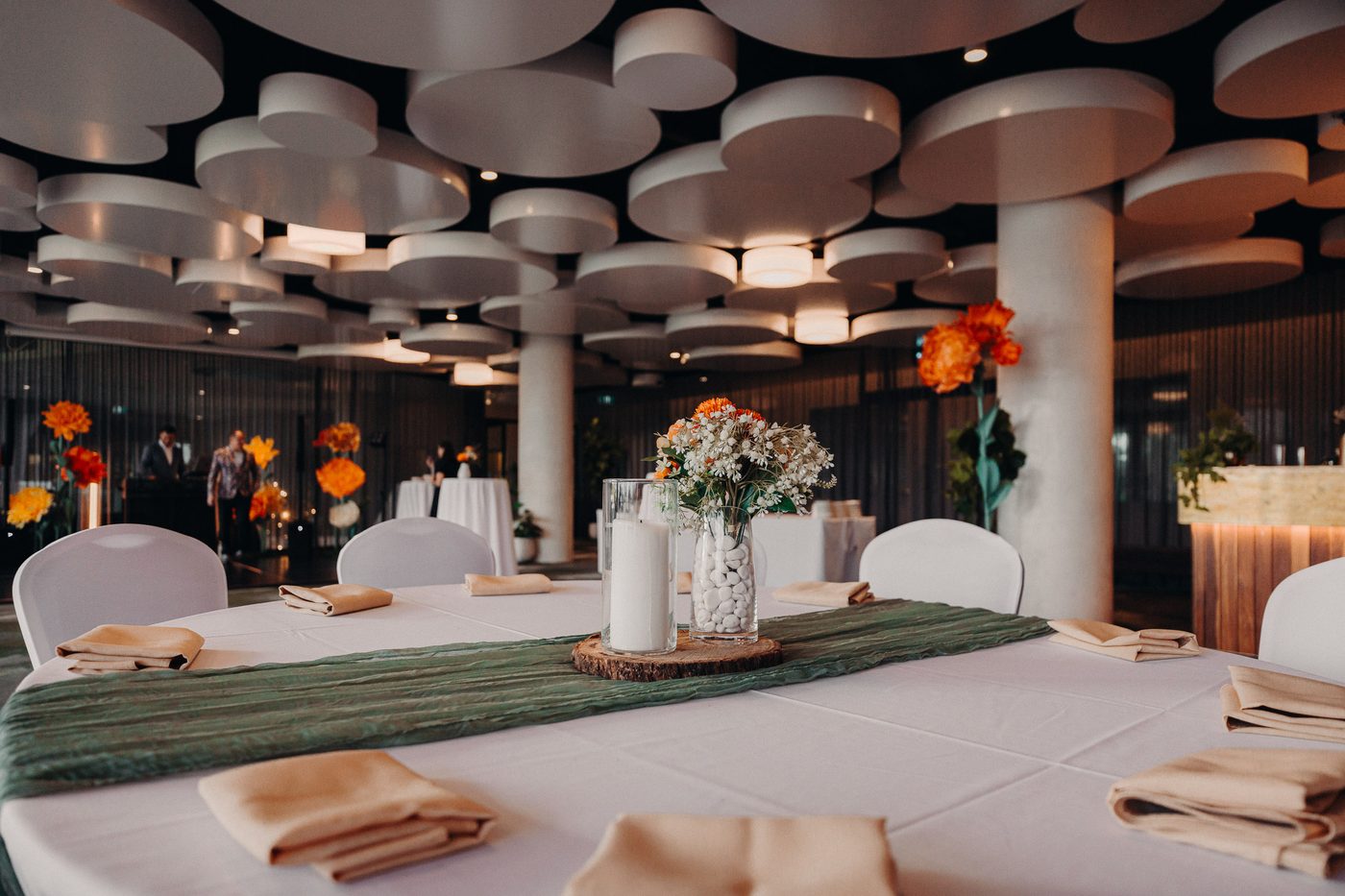 Round table set with beige napkins, a green table runner, and a centerpiece of white flowers and candles, with guests and floral decorations visible in the background. Wedding fair season Brisbane by She Said Yes.