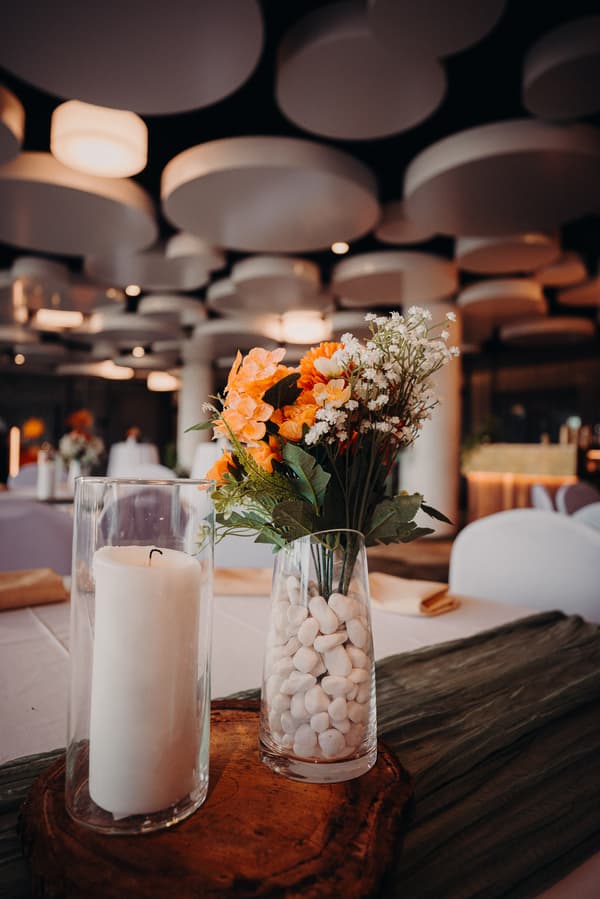 A floral centerpiece with orange and white flowers in a glass vase filled with white stones, alongside a large white candle in a glass holder, set on a wooden slab on a table at Eatons Hill Hotel — Hills Room.