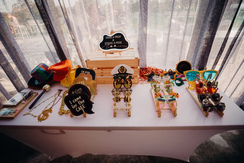 A table at Eatons Hill Hotel — Hills Room is set up with various wedding photo booth props including hats, oversized glasses, novelty necklaces, and speech bubble signs with phrases like 'I came for the CAKE' and 'GRAB A PROP & STRIKE A POSE'.