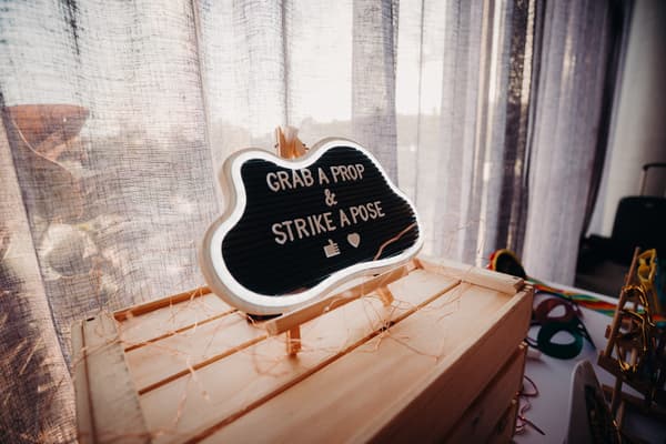 A sign on a wooden crate at Eatons Hill Hotel — Hills Room reads 'GRAB A PROP & STRIKE A POSE' with a thumbs-up and heart symbol, surrounded by string lights and photo booth props on a table.