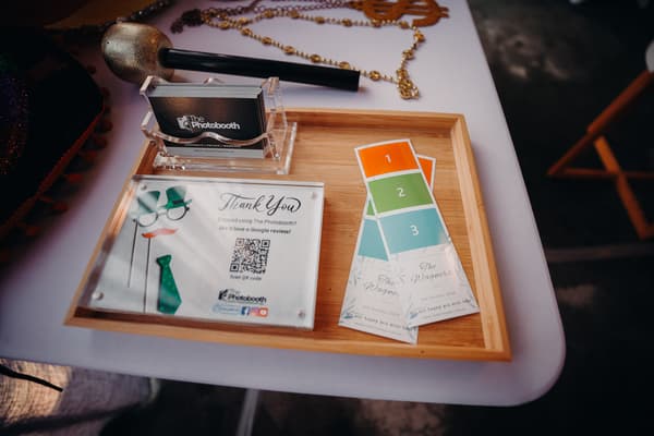 A wooden tray on a white table at Eatons Hill Hotel — Hills Room holds a microphone, a clear stand with The Photobooth business cards, a thank you sign with a QR code for reviews, and two photo strip prints labeled 'The Wagner's' dated 6th October 2024.