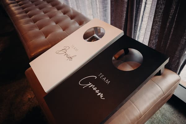 Two cornhole boards labeled 'Team Bride' and 'Team Groom' resting on a padded bench near a window with curtains at Eatons Hill Hotel — Hills Room.