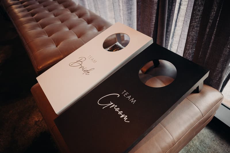 Two cornhole boards labeled 'Team Bride' and 'Team Groom' resting on a padded bench near a window with curtains at Eatons Hill Hotel — Hills Room.