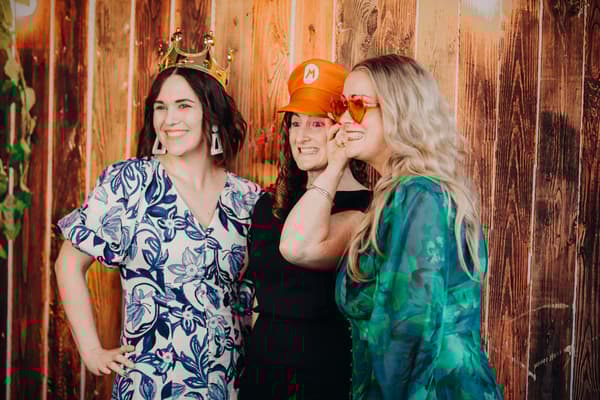 Three female guests pose together wearing playful accessories against a wooden wall at Eatons Hill Hotel — Hills Room.