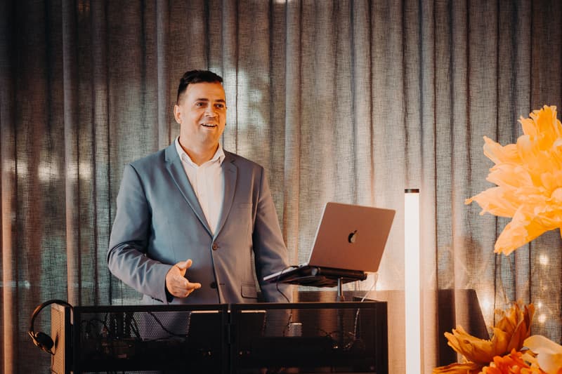 A man in a suit stands behind a DJ booth with a laptop at Eatons Hill Hotel — Hills Room during the wedding reception.