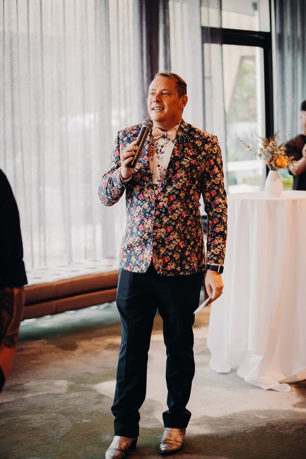 A man in a floral jacket and bow tie holds a microphone and a note card while speaking at the reception in the Hills Room at Eatons Hill Hotel.