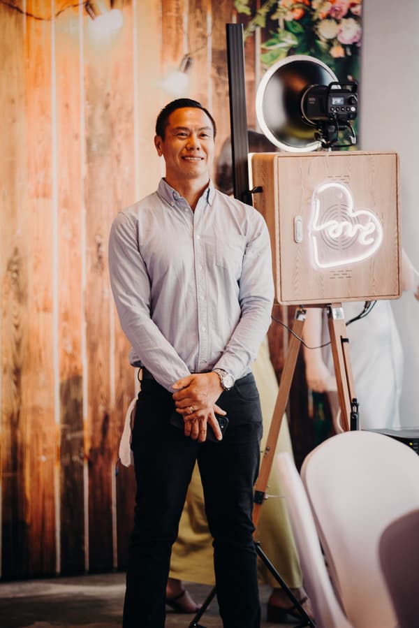 A man in a light blue button-up shirt and black pants stands near a wooden photo booth with a neon heart-shaped 'love' sign at Eatons Hill Hotel — Hills Room during the wedding reception.