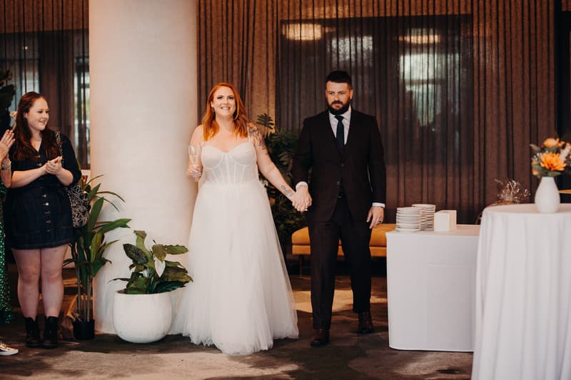 The bride Lilas in a white wedding gown holding a glass and the groom Kaine in a dark suit holding hands while entering the reception area at Eatons Hill Hotel — Hills Room. Guests stand nearby clapping.