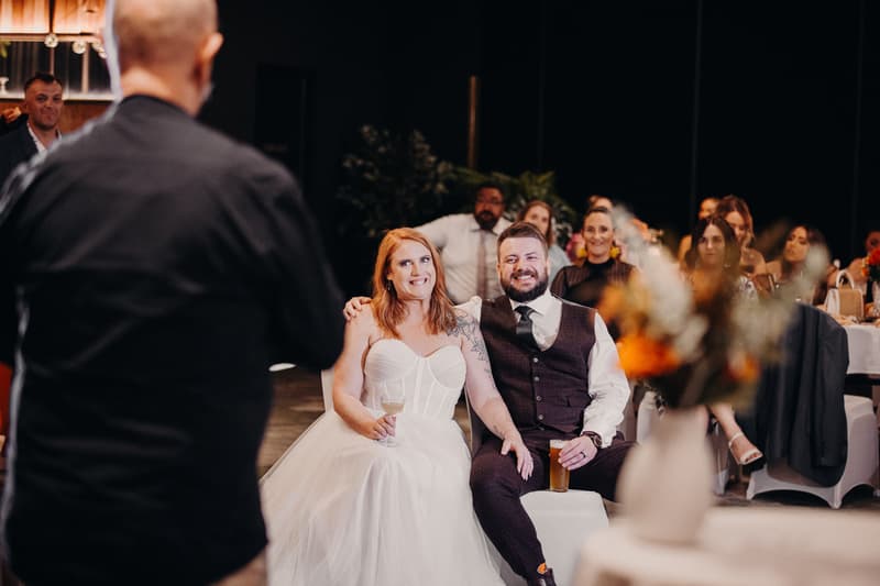 Bride Lilas and groom Kaine sit together at the Eatons Hill Hotel — Hills Room reception, holding drinks and facing a speaker addressing the guests.