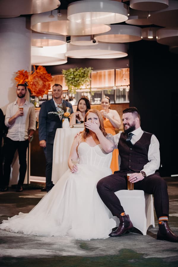 The bride Lilas in a white wedding gown sits next to the groom Kaine in a dark suit and vest at Eatons Hill Hotel — Hills Room during the reception. Lilas covers her mouth with her hand while holding a glass, and Kaine has his arm around her shoulder, holding a drink. Behind them, guests stand near a high table with floral arrangements and drinks.