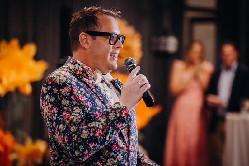A man wearing a floral jacket and glasses holds a microphone and speaks at the reception stage in the Hills Room at Eatons Hill Hotel, with two blurred guests in the background.