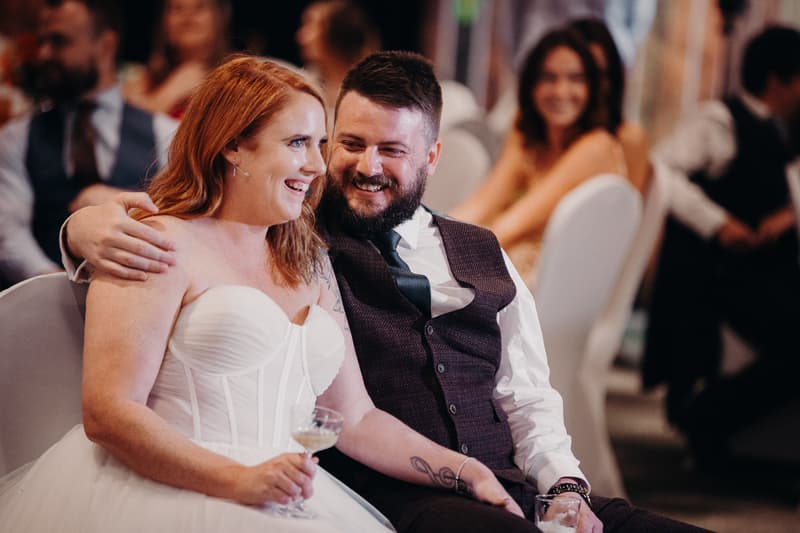The bride Lilas and groom Kaine sit closely together at the reception in the Hills Room at Eatons Hill Hotel, with Lilas holding a glass and Kaine's arm around her shoulder. Guests are seated in the background.