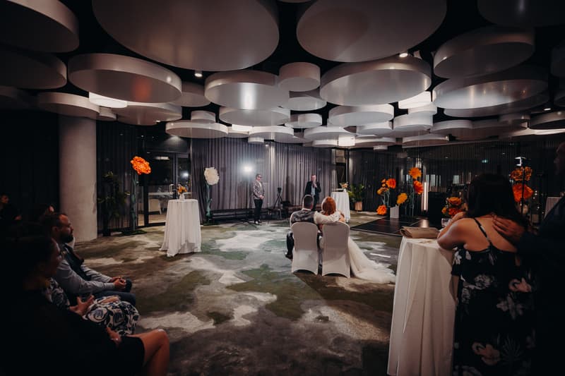 Bride Lilas and groom Kaine sit side by side on white chairs facing a speaker at the Eatons Hill Hotel Hills Room during the wedding reception. Guests are seated and standing around, with floral arrangements and round ceiling decorations visible.