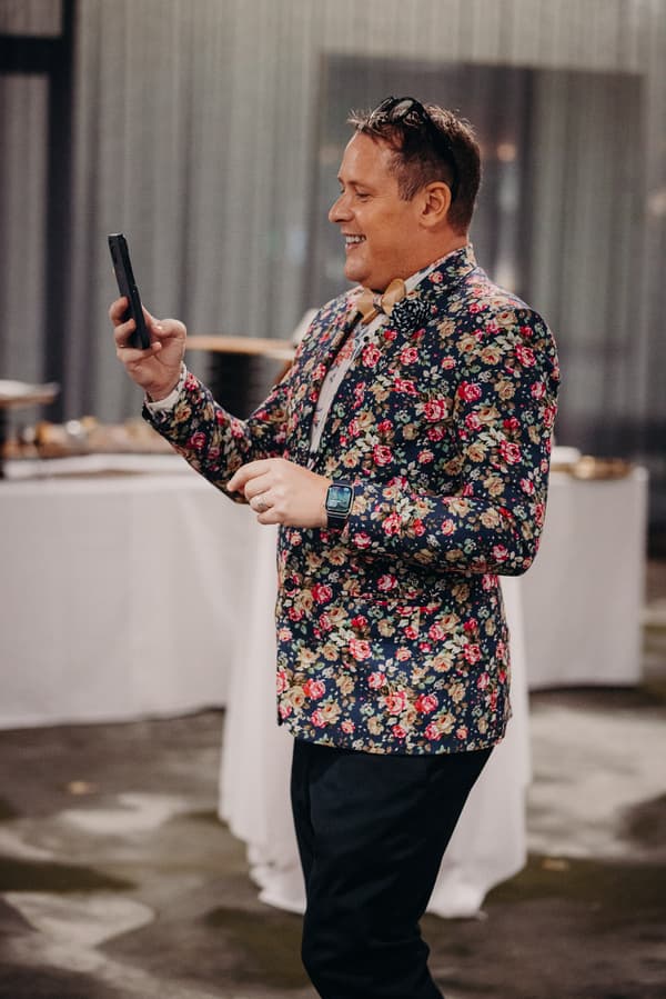 A male guest wearing a floral jacket and bow tie is holding a smartphone and smiling at the reception in the Hills Room at Eatons Hill Hotel.
