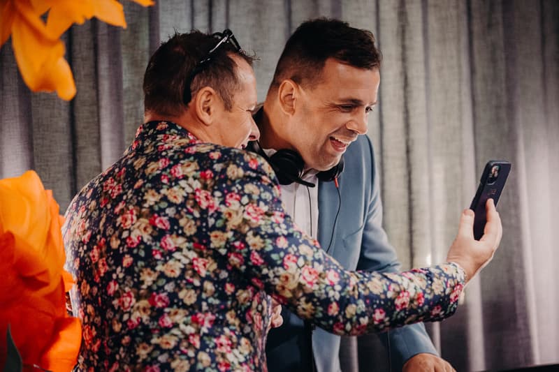Two men at the Eatons Hill Hotel Hills Room reception stage, one wearing a floral jacket and the other in a blue suit with headphones around his neck, looking at a smartphone together.