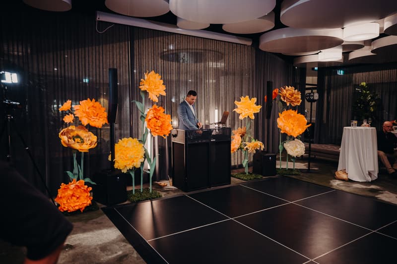 A DJ stands behind a console surrounded by large orange and yellow floral decorations next to a black dance floor at Eatons Hill Hotel — Hills Room during the wedding reception.