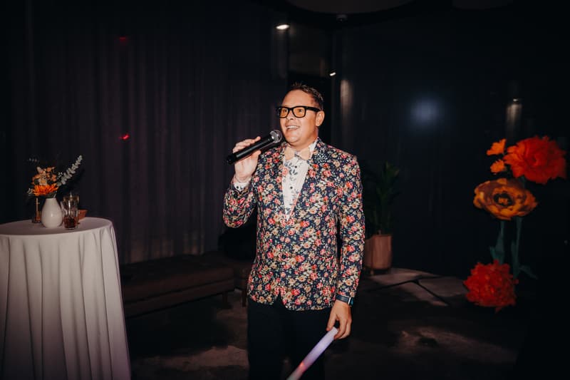 A man in a floral jacket and glasses holds a microphone and appears to be speaking at the reception stage in the Hills Room at Eatons Hill Hotel. There is a tall round table with a vase of flowers and drinks to his left, and large decorative orange flowers in the background.