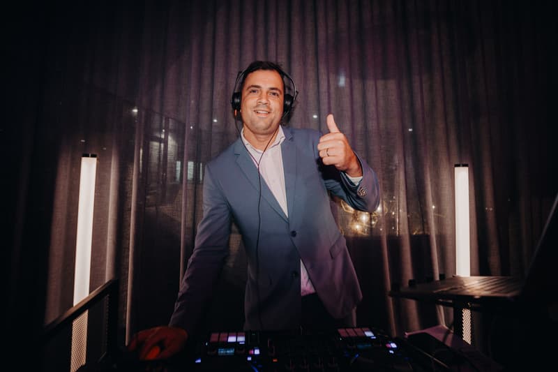 A man wearing headphones and a suit stands behind DJ equipment giving a thumbs-up gesture at the Eatons Hill Hotel — Hills Room during the wedding reception.