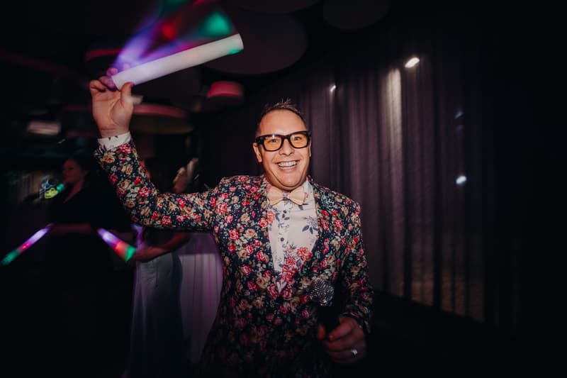 A male guest wearing a floral jacket and bow tie holds a microphone and a glowing stick at the reception in the Hills Room at Eatons Hill Hotel.