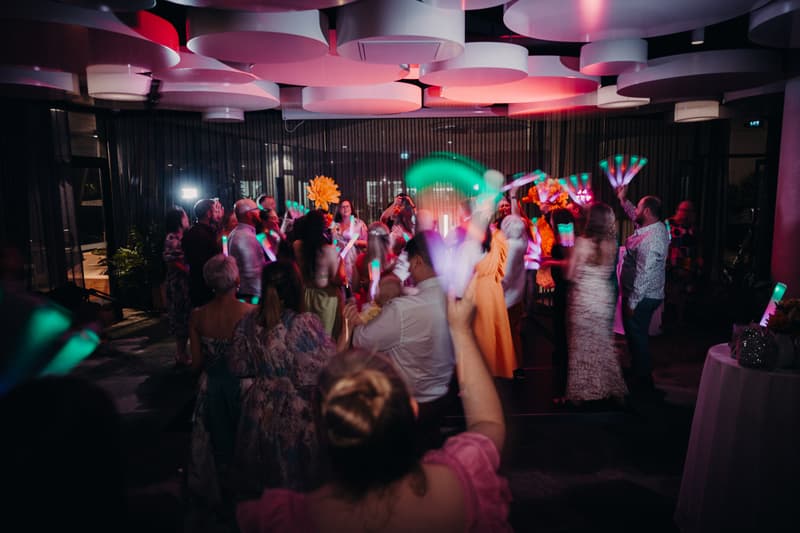 Guests dance and celebrate at the reception in the Hills Room at Eatons Hill Hotel, holding light-up sticks and fans under circular ceiling fixtures.