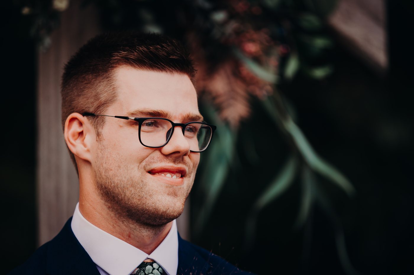 Connor, the groom, wearing glasses and a dark suit with a patterned tie, stands near a wooden post with blurred greenery in the background. Lead follow-up communication issues by She Said Yes.