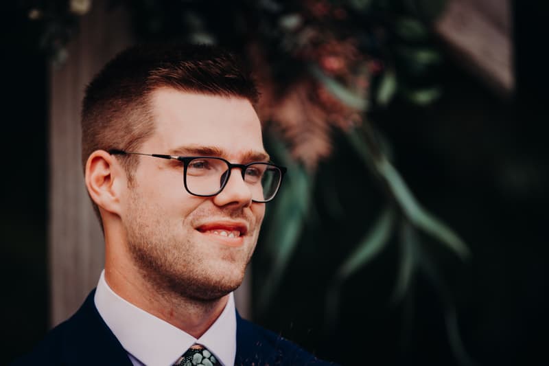 Connor, the groom, wearing glasses and a dark suit with a patterned tie, stands near a wooden post with blurred greenery in the background at Yabbaloumba Retreat — By The River.