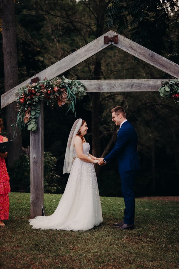 Bride Lilly and groom Connor hold hands facing each other under a wooden arch decorated with greenery and flowers at Yabbaloumba Retreat — By The River during their wedding ceremony.