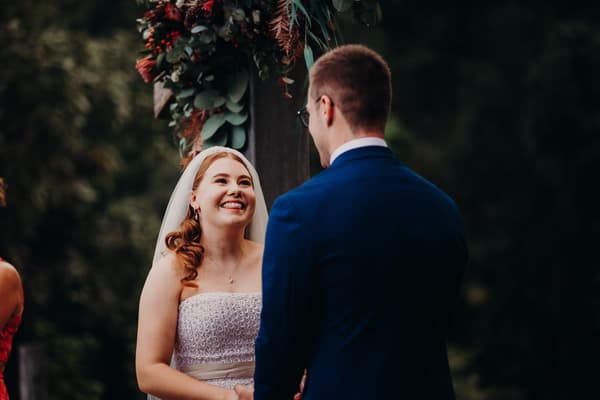 Bride Lilly and groom Connor stand facing each other under a floral arch at Yabbaloumba Retreat — By The River during their wedding ceremony.