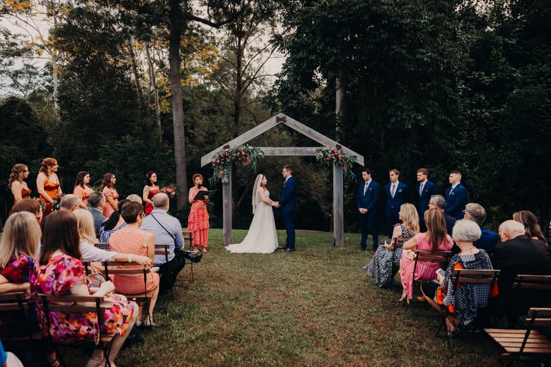 Bride Lilly and groom Connor stand holding hands under a wooden arch decorated with flowers at Yabbaloumba Retreat — By The River, while an officiant in a red dress reads from a book. Bridesmaids in orange dresses stand to the left, groomsmen in blue suits stand to the right, and guests seated on wooden chairs watch the ceremony.
