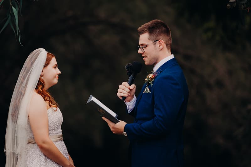 Bride Lilly and groom Connor stand facing each other at the ceremony stage at Yabbaloumba Retreat — By The River. Connor is holding a microphone and reading from a book, while Lilly listens attentively wearing her wedding dress and veil.