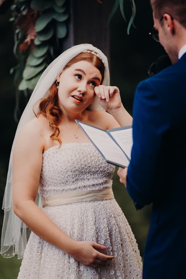 The bride Lilly wipes a tear while the groom Connor reads from a booklet during their wedding ceremony at Yabbaloumba Retreat — By The River.