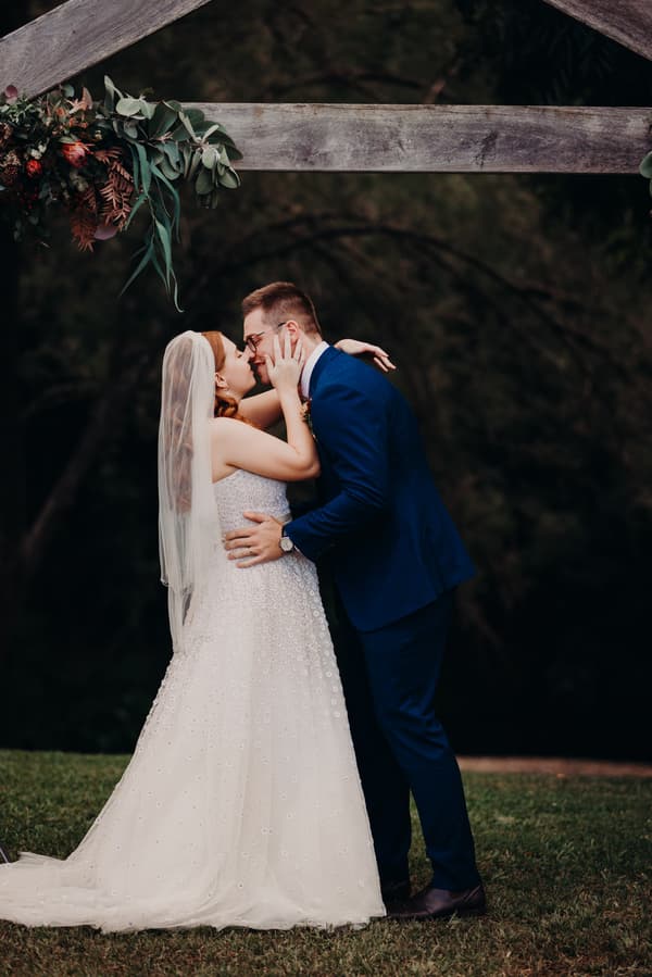 Bride Lilly and groom Connor embrace and kiss under a wooden arch decorated with greenery and flowers at Yabbaloumba Retreat — By The River.