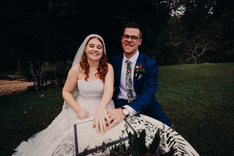 Bride Lilly and groom Connor sit together at a white ornate table outdoors on grass at Yabbaloumba Retreat — By The River, showing their wedding rings and a signed document.
