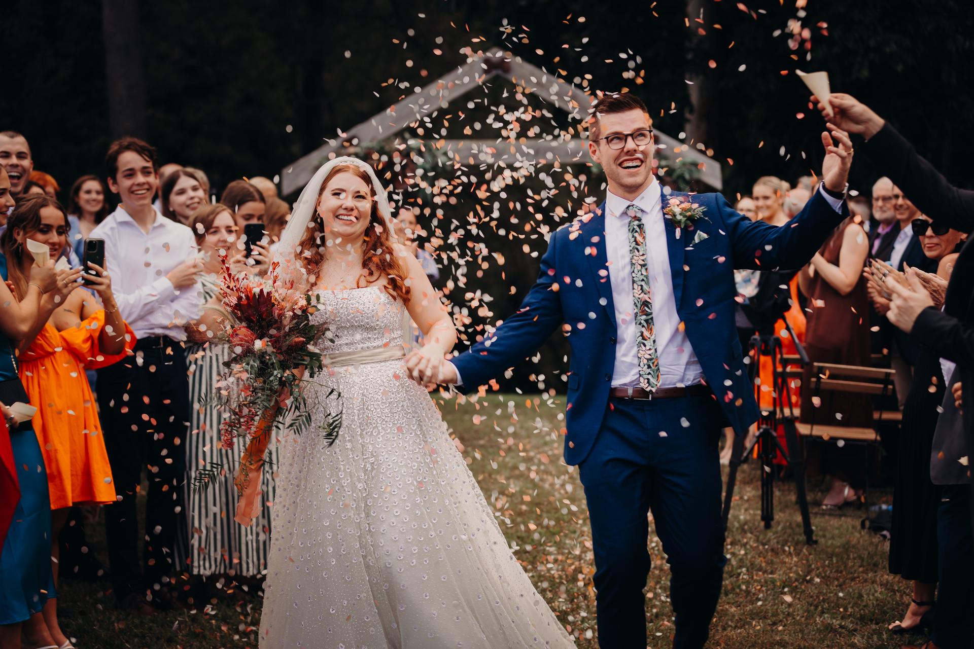 Bride Lilly and groom Connor walk down the aisle at Yabbaloumba Retreat — By The River while guests throw confetti.