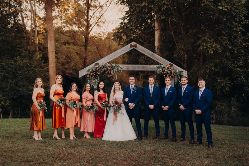 Lilly the bride and Connor the groom stand together with their bridesmaids and groomsmen under a wooden arch decorated with floral arrangements at Yabbaloumba Retreat.
