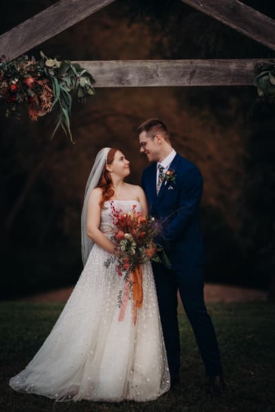 Bride Lilly and groom Connor stand together under a wooden arch decorated with greenery and flowers at Yabbaloumba Retreat. Lilly wears a strapless white wedding gown with a veil and holds a bouquet of flowers with orange and red tones. Connor wears a dark blue suit with a floral tie and boutonniere. They look at each other while standing on grass with a blurred natural background.