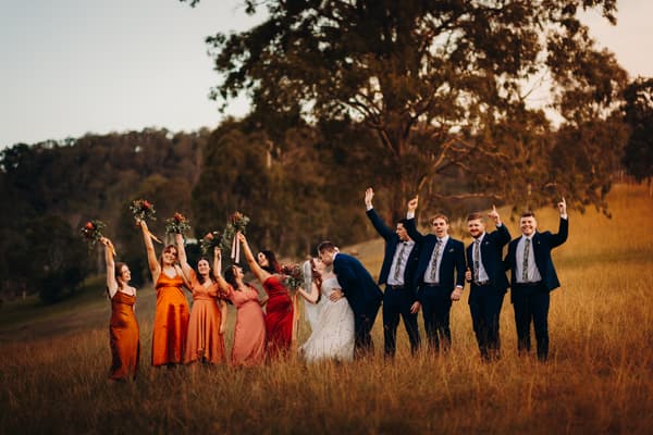 Bride Lilly and groom Connor kiss in a grassy field at Yabbaloumba Retreat, surrounded by bridesmaids in orange and red dresses holding bouquets and groomsmen in navy suits with floral ties raising their hands.