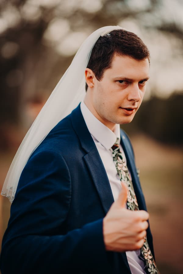 Connor, the groom, wearing a dark blue suit and floral tie, poses outdoors at Yabbaloumba Retreat with a white bridal veil on his head, giving a thumbs-up gesture.