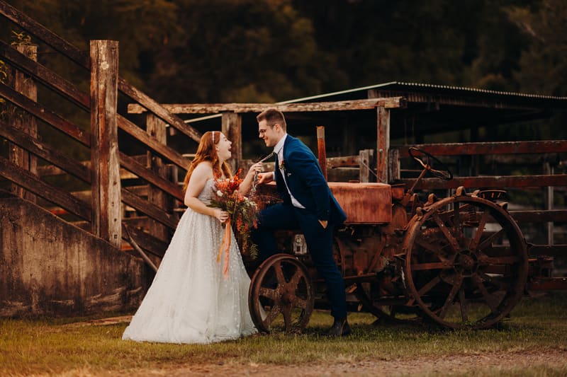 Bride Lilly in a white wedding gown holding a bouquet and groom Connor in a blue suit pose together beside a rusty old tractor at Yabbaloumba Retreat.
