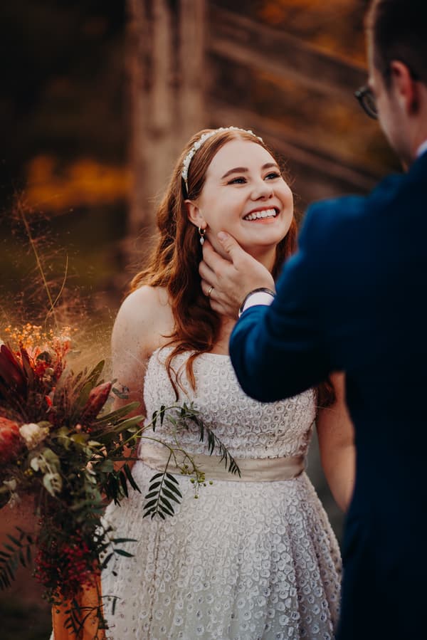 The bride Lilly smiles as the groom Connor gently holds her face during their couple portraits at Yabbaloumba Retreat. Lilly is wearing a white textured wedding dress and holding a bouquet with greenery and red flowers. Connor is dressed in a blue suit.