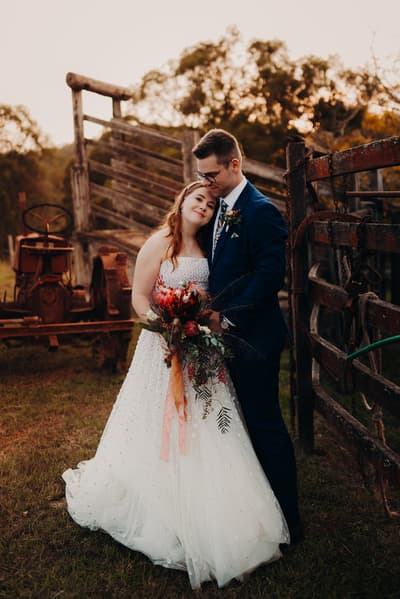 Bride Lilly in a white wedding gown holding a bouquet and groom Connor in a navy suit embrace outdoors near rustic wooden fencing and an old tractor at Yabbaloumba Retreat.