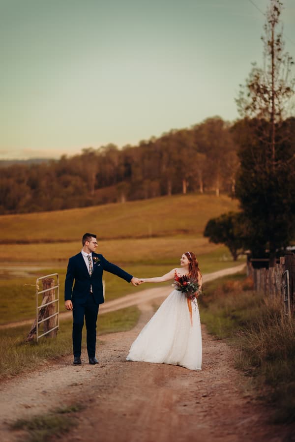 Bride Lilly and groom Connor hold hands on a dirt path at Yabbaloumba Retreat, surrounded by grassy fields and trees in the background during their couple portraits session.