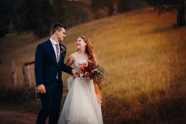 Bride Lilly and groom Connor walk hand in hand outdoors at Yabbaloumba Retreat, with Lilly holding a bouquet of flowers and wearing a strapless wedding dress, and Connor in a dark suit with a floral tie.
