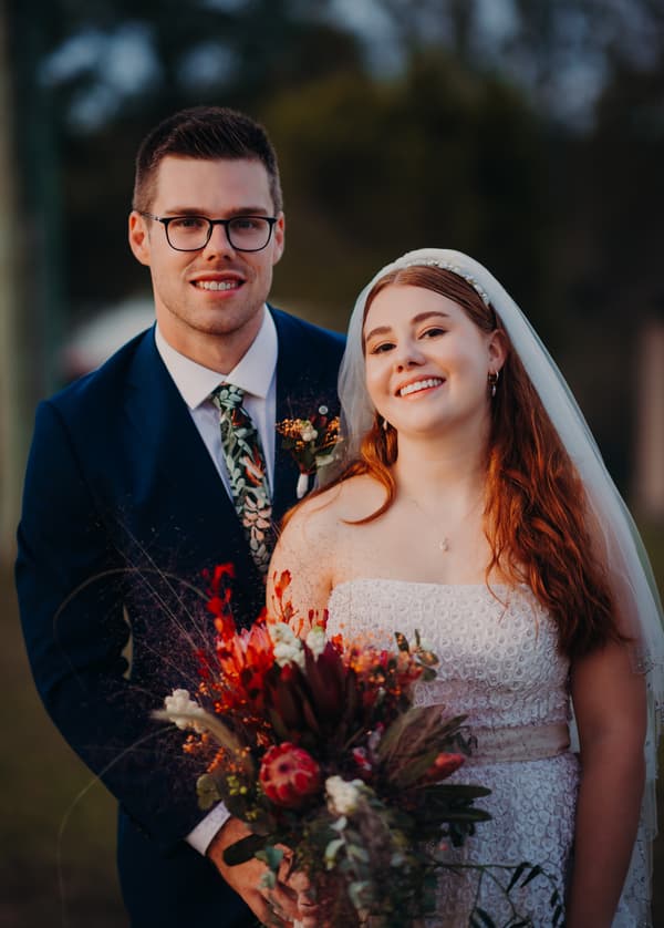 Bride Lilly and groom Connor pose together outdoors at Yabbaloumba Retreat, with Lilly holding a colorful bouquet and wearing a strapless white wedding dress and veil, and Connor in a dark blue suit with a floral tie and boutonniere.